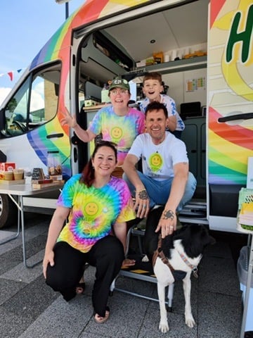 Four smiling people and a dog posing at the doorway of a colorful rainbow-decorated food truck or van