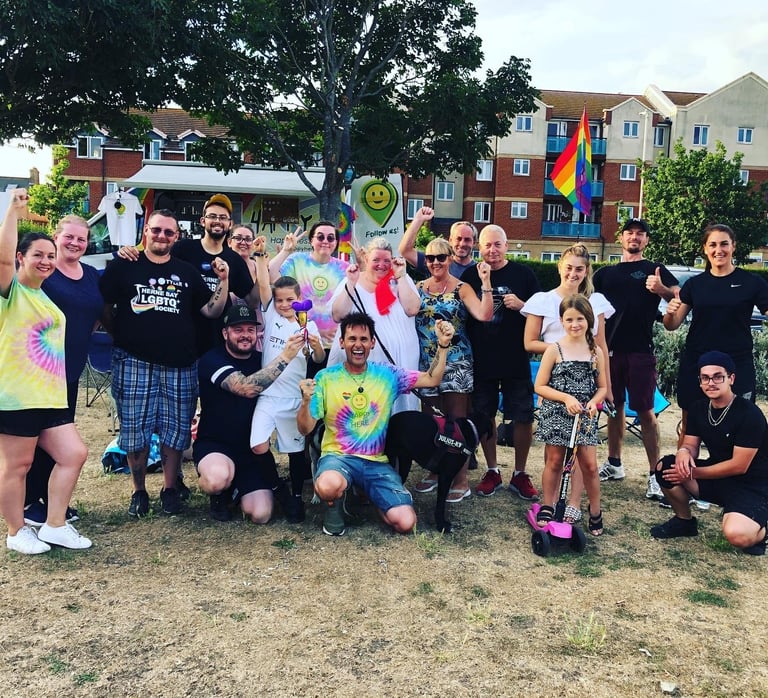 Large group of adults and children posing together outdoors at a community event near colorful buildings and a rainbow flag