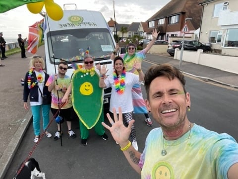 Man in colorful tie-dye shirt takes selfie with children and decorated parade vehicle on residential street